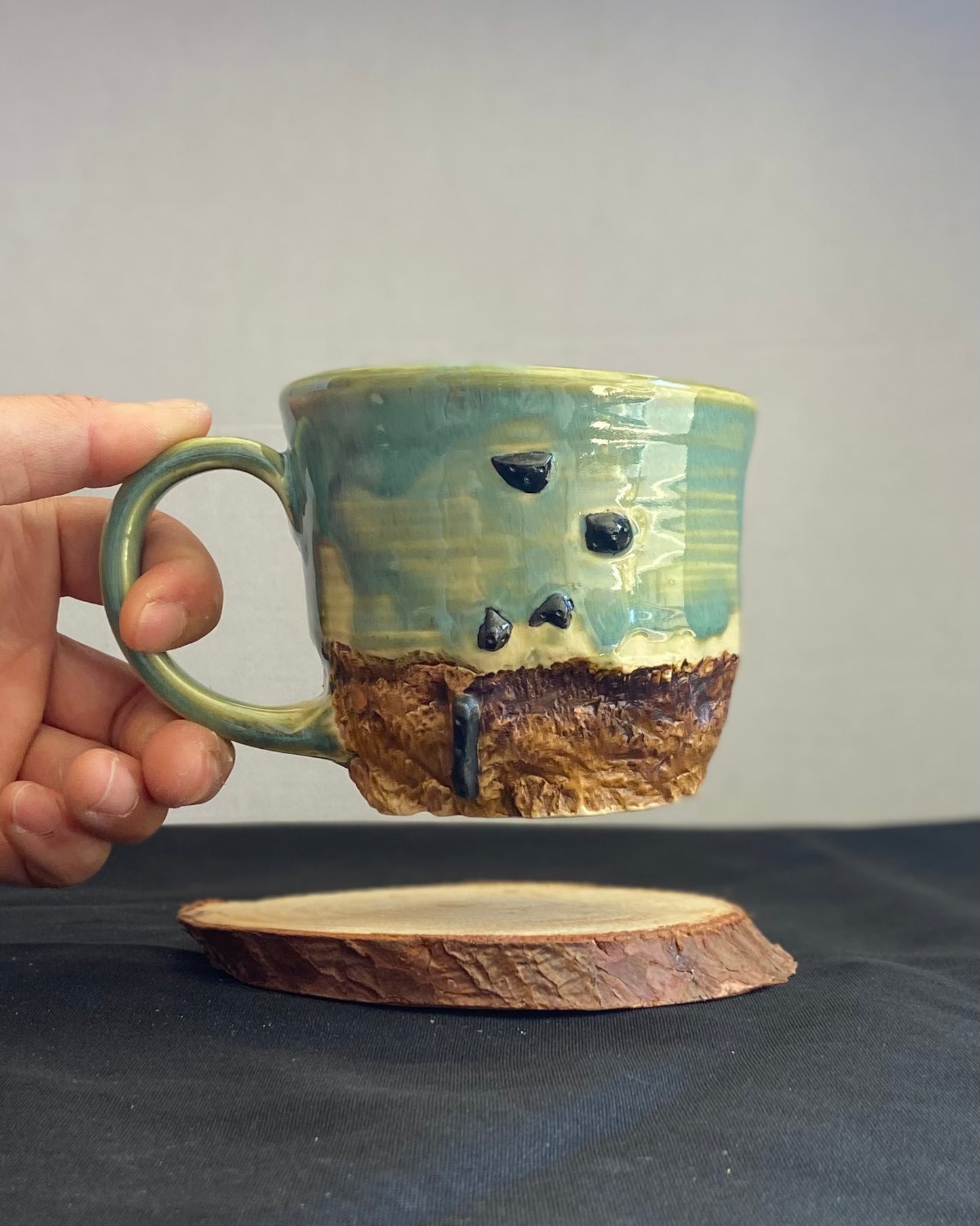 Handcrafted Boulder Mug with glossy celadon sky glaze and rocky textured mountainscape, featuring miniature black rock climbing holds with tiny bolt holes. Unique ceramic mug inspired by indoor and outdoor rock climbing adventures, perfect for climbers. Artisan quality, wabi sabi design, and playful glaze details.