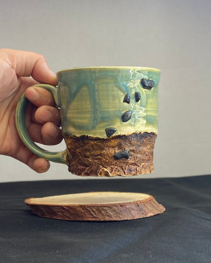 Handcrafted Boulder Mug with glossy celadon sky glaze and rocky textured mountainscape, featuring miniature black rock climbing holds with tiny bolt holes. Unique ceramic mug inspired by indoor and outdoor rock climbing adventures, perfect for climbers. Artisan quality, wabi sabi design, and playful glaze details.