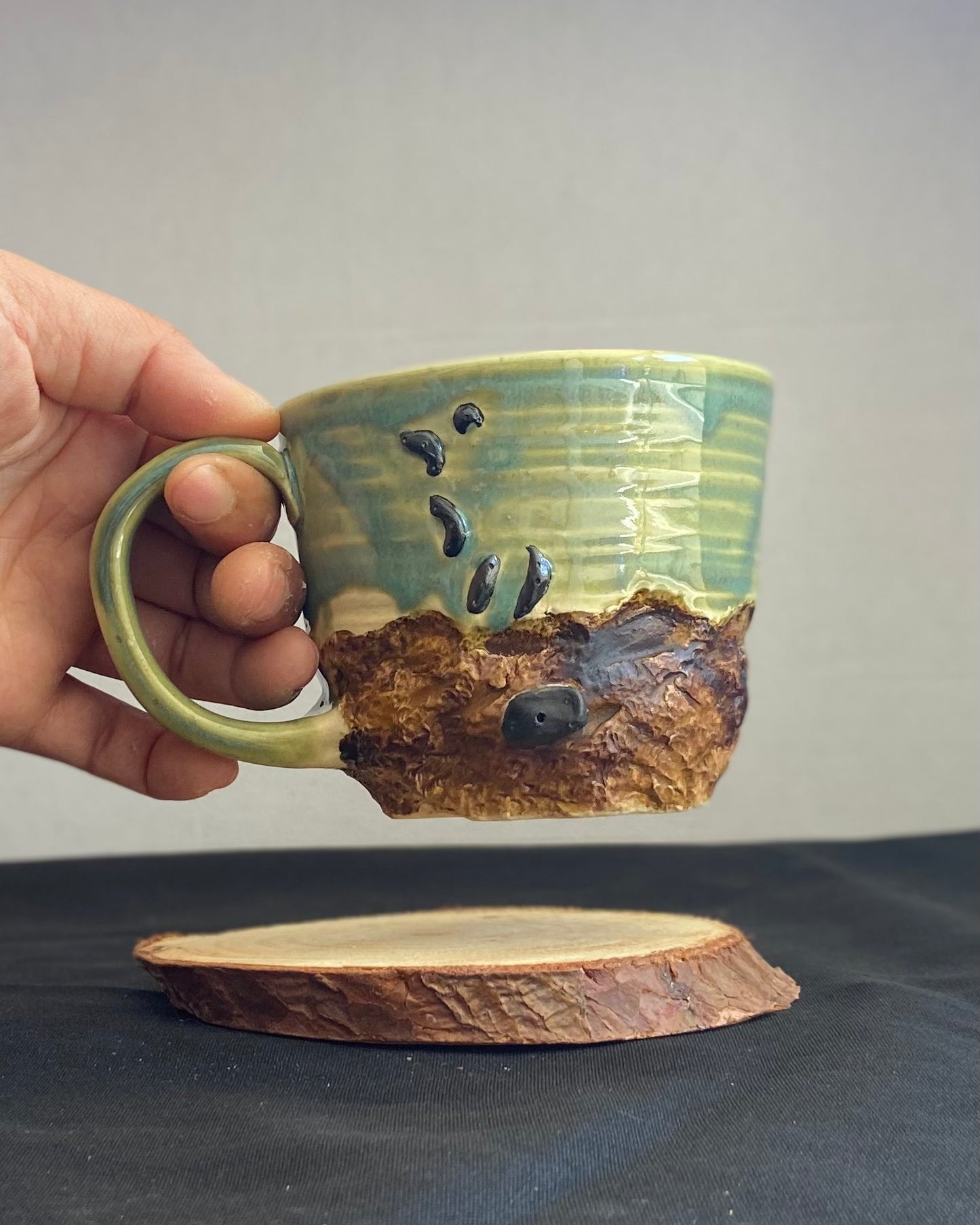 Handcrafted Boulder Mug with glossy celadon sky glaze and rocky textured mountainscape, featuring miniature black rock climbing holds with tiny bolt holes. Unique ceramic mug inspired by indoor and outdoor rock climbing adventures, perfect for climbers. Artisan quality, wabi sabi design, and playful glaze details.