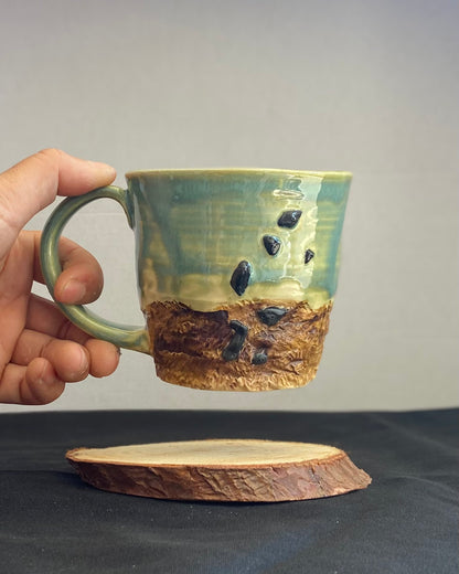 Handcrafted Boulder Mug with glossy celadon sky glaze and rocky textured mountainscape, featuring miniature black rock climbing holds with tiny bolt holes. Unique ceramic mug inspired by indoor and outdoor rock climbing adventures, perfect for climbers. Artisan quality, wabi sabi design, and playful glaze details.