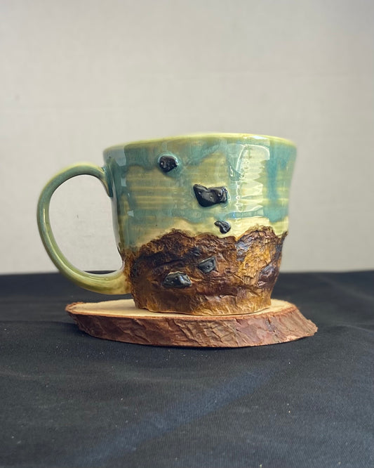 Handcrafted Boulder Mug with glossy celadon sky glaze and rocky textured mountainscape, featuring miniature black rock climbing holds with tiny bolt holes. Unique ceramic mug inspired by indoor and outdoor rock climbing adventures, perfect for climbers. Artisan quality, wabi sabi design, and playful glaze details.