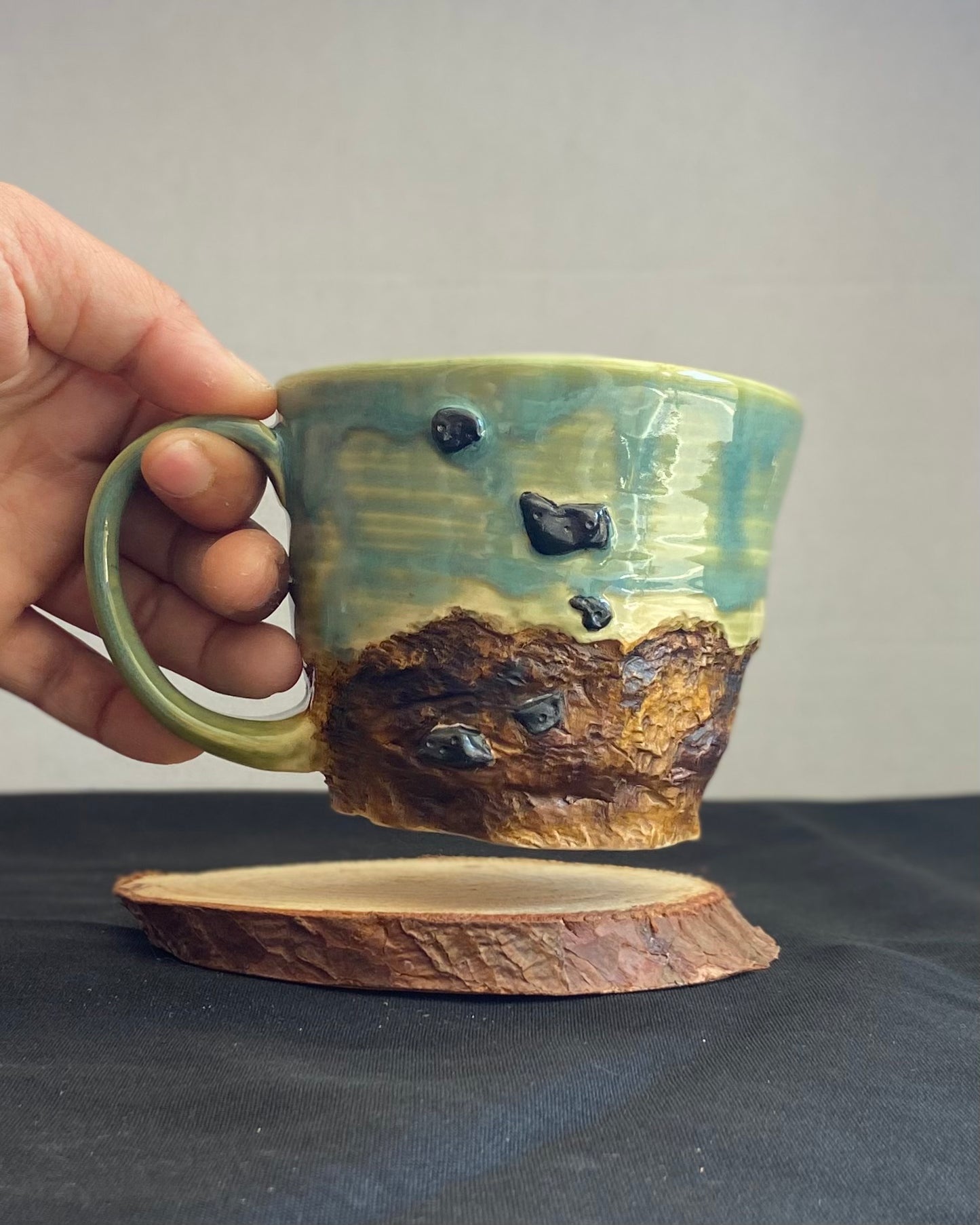 Handcrafted Boulder Mug with glossy celadon sky glaze and rocky textured mountainscape, featuring miniature black rock climbing holds with tiny bolt holes. Unique ceramic mug inspired by indoor and outdoor rock climbing adventures, perfect for climbers. Artisan quality, wabi sabi design, and playful glaze details.