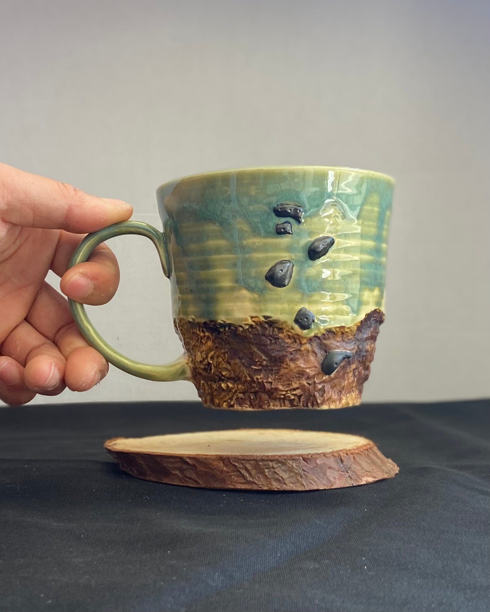 Handcrafted Boulder Mug with glossy celadon sky glaze and rocky textured mountainscape, featuring miniature black rock climbing holds with tiny bolt holes. Unique ceramic mug inspired by indoor and outdoor rock climbing adventures, perfect for climbers. Artisan quality, wabi sabi design, and playful glaze details. Hand-etched joshua tree.