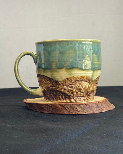 Handcrafted Mountain Mug with glossy celadon sky glaze and rocky textured mountainscape, accented with iron oxide and matte amber quartz glaze. Unique ceramic mug inspired by outdoor rock climbing, perfect for coffee or tea. Artisan quality, wabi sabi design, and playful glaze details