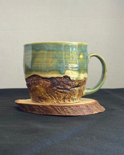 Handcrafted Mountain Mug with glossy celadon sky glaze and rocky textured mountainscape, accented with iron oxide and matte amber quartz glaze. Unique ceramic mug inspired by outdoor rock climbing, perfect for coffee or tea. Artisan quality, wabi sabi design, and playful glaze details