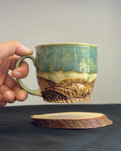 Handcrafted Mountain Mug with glossy celadon sky glaze and rocky textured mountainscape, accented with iron oxide and matte amber quartz glaze. Unique ceramic mug inspired by outdoor rock climbing, perfect for coffee or tea. Artisan quality, wabi sabi design, and playful glaze details