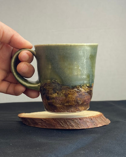 Handthrown ceramic mug, sage matte glazed with amber-iron oxide wash. Dual-texture surface, matte. Rock-hammered rugged mountain landscape, hand-pulled handle. Fired to cone 5.