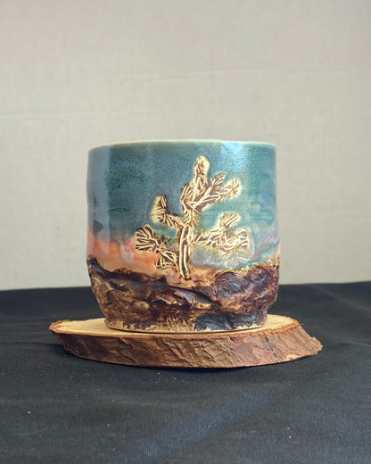 Handcrafted tumbler with hand-etched Joshua tree, antique blue satin glaze, and sunburnt pink glaze creating a sunset over a hand-carved mountain landscape. Unique ceramic tumbler inspired by nature, perfect for beverages. Artisan quality, wabi sabi design, and nature-inspired aesthetic