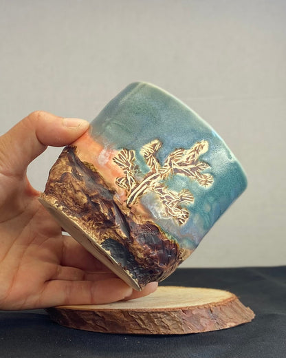 Handcrafted tumbler with hand-etched Joshua tree, antique blue satin glaze, and sunburnt pink glaze creating a sunset over a hand-carved mountain landscape. Unique ceramic tumbler inspired by nature, perfect for beverages. Artisan quality, wabi sabi design, and nature-inspired aesthetic