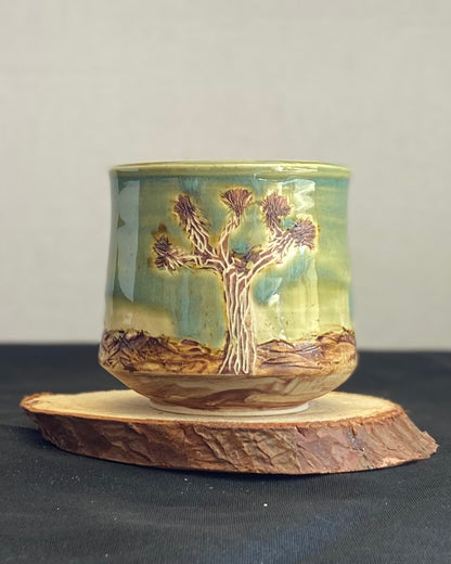 Handcrafted tumbler with hand-etched Joshua tree, celadon blue-green glaze over a hand-carved mountain landscape. Unique ceramic tumbler inspired by nature, perfect for beverages. Artisan quality, wabi sabi design, and nature-inspired aesthetic