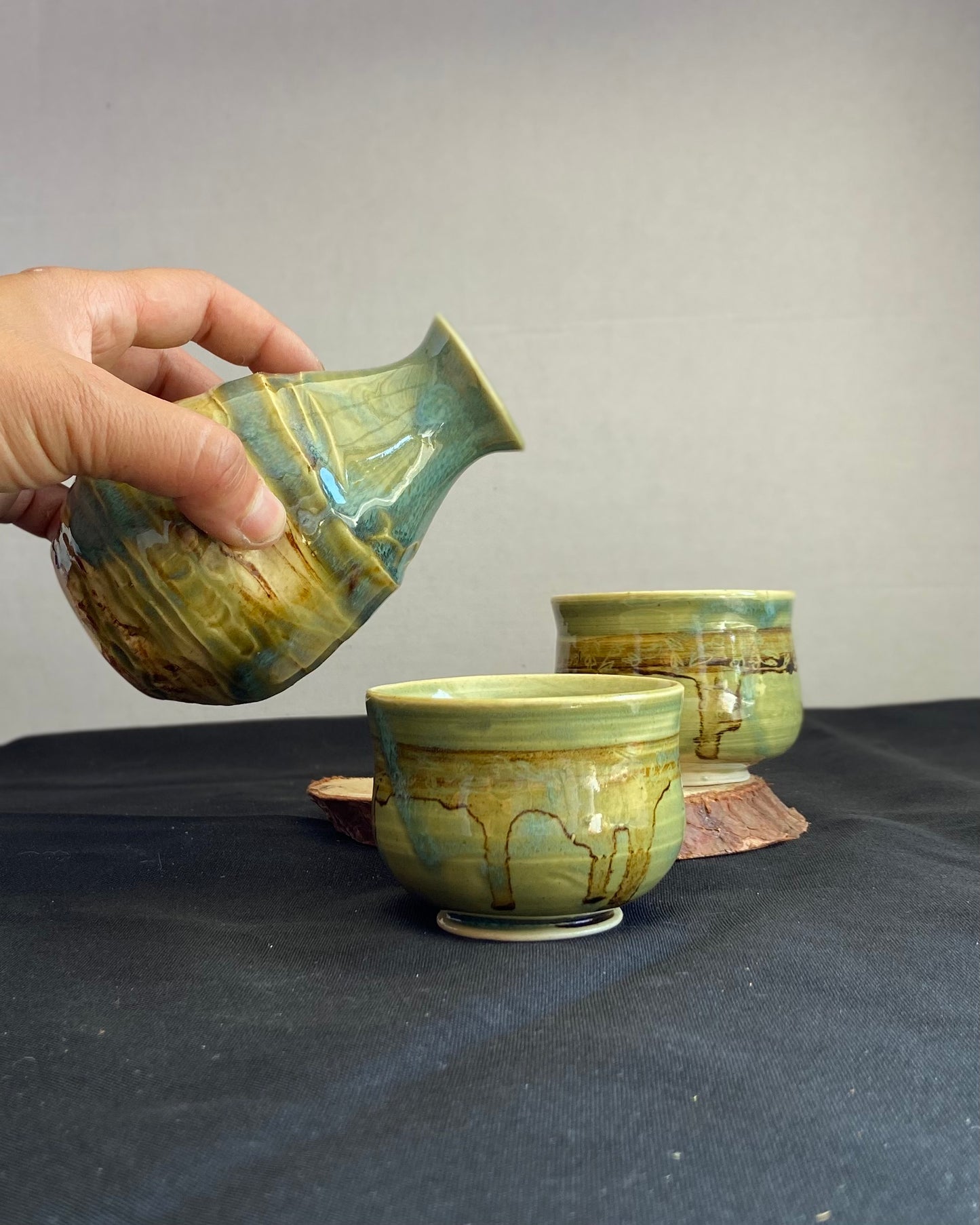 Handcrafted sake set with one pourer and two cups, featuring celadon glaze over textured carvings to evoke water. Cups have dripping iron oxide accents and tulips carved on the lip. Unique ceramic sake set inspired by nature, perfect for serving sake. Artisan quality, wabi sabi design, and nature-inspired aesthetic